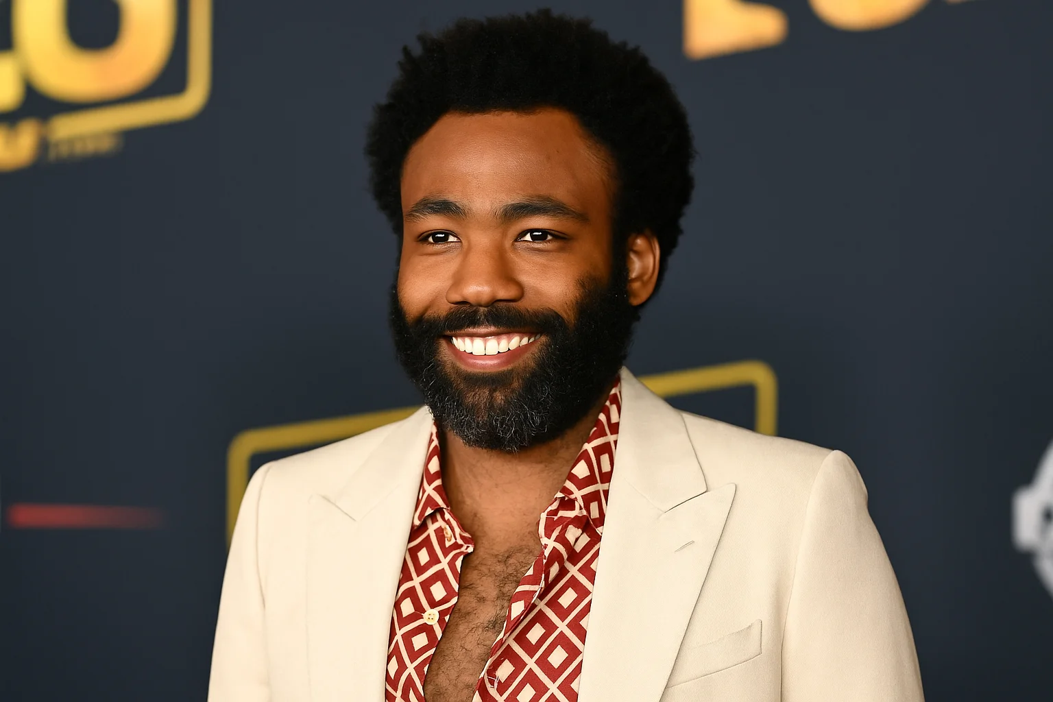 Donald Glover smiling at a red carpet event, wearing a cream suit jacket and patterned red shirt.