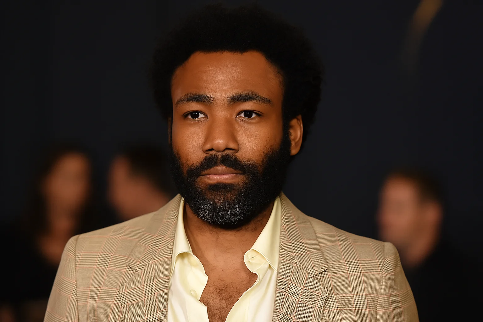 Donald Glover wearing a beige plaid suit jacket and yellow shirt, looking composed at a formal event.